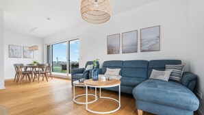 Ein modernes Wohnzimmer mit einem blauen Ecksofa, einem Essbereich mit Holzsesseln und einem großen Fenster mit Blick ins Freie.