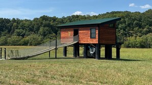 A wooden cabin with a green roof, elevated on stilts, featuring a small porch and a rope bridge leading to it. - Tarzan And Jane Treehouse. Hot tub. Riverfront (Sevierville)