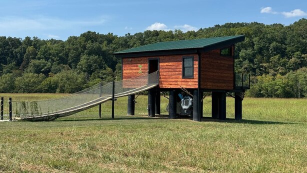 A wooden cabin with a green roof, elevated on stilts, featuring a small porch and a rope bridge leading to it.