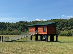 Una cabaña de madera con techo verde, elevada sobre pilotes, que cuenta con un pequeño porche y un puente de cuerda que la conecta.
