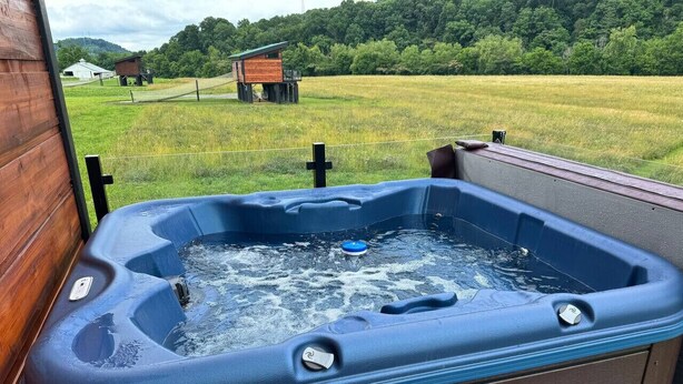 A hot tub with a wooden exterior, set on a porch with a view of a grassy field and a small wooden structure in the background. - Tarzan And Jane Treehouse. Hot tub. Riverfront (Sevierville)