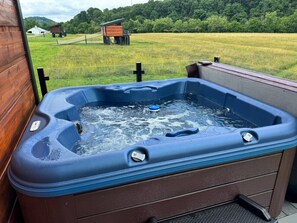 A hot tub with a wooden exterior, set on a porch with a view of a grassy field and a small wooden structure in the background. - Tarzan And Jane Treehouse. Hot tub. Riverfront (Sevierville)