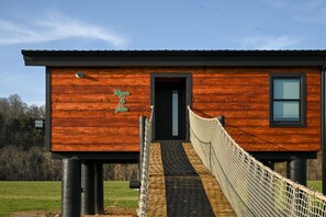 Une cabane moderne en bois, accessible par un pont de corde qui mène à l’entrée.