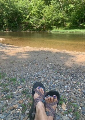 A pair of feet in sandals on a rocky riverbank with a river and greenery in the background.