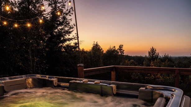 Een hottubgedeelte met een houten terras en lichtjes, omringd door bomen en uitzicht op een zonsondergang.