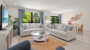 Ein modernes Wohnzimmer mit einem Ecksofa, einem Glastisch, einem Essbereich mit Tisch und Stühlen sowie großen Fenstern mit Vorhängen.