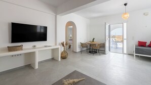 A modern living room with a flat-screen TV, a bench, a dining area with chairs, and a view through an open door.
