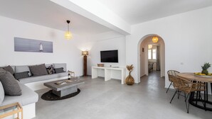 A modern living room with a grey sofa, a flat-screen TV, a coffee table, and a dining area with wicker chairs.