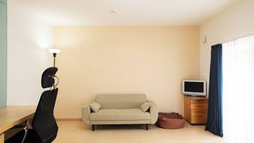 A minimalist living room with a sofa, a TV on a wooden cabinet, a desk with a chair, and a window with curtains.