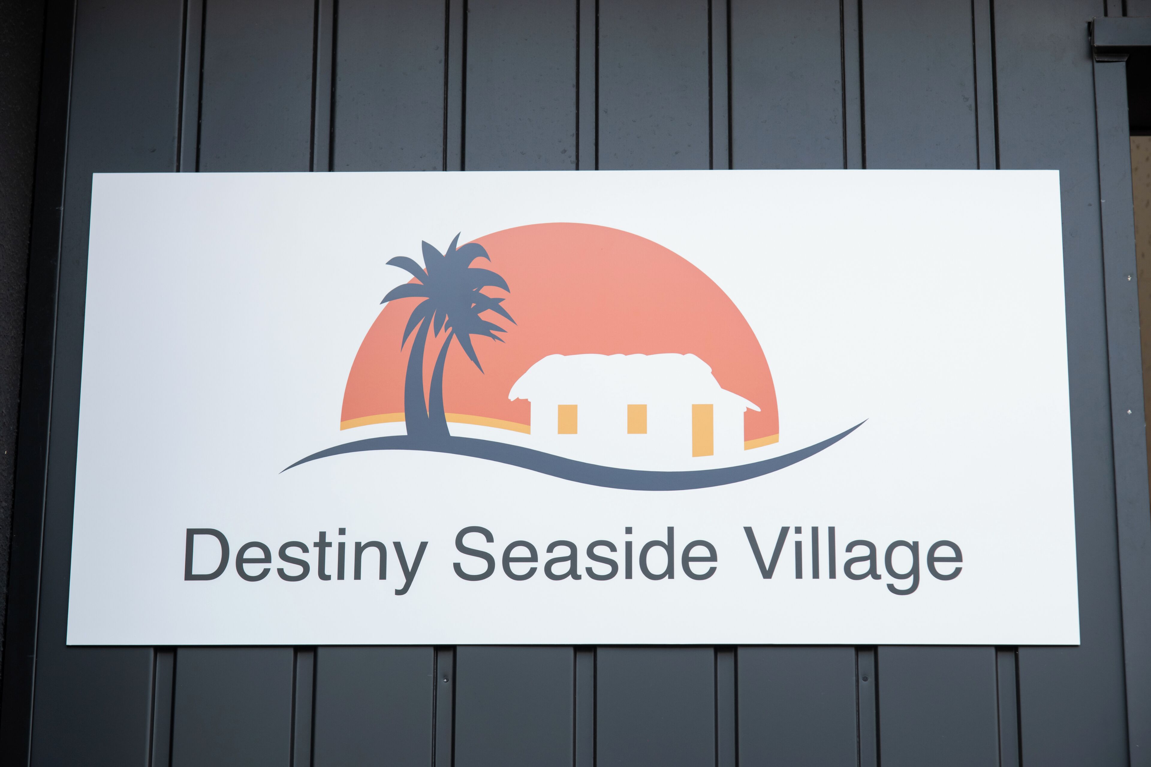 A sign for Destiny Seaside Village with a palm tree and a house against a sunset.