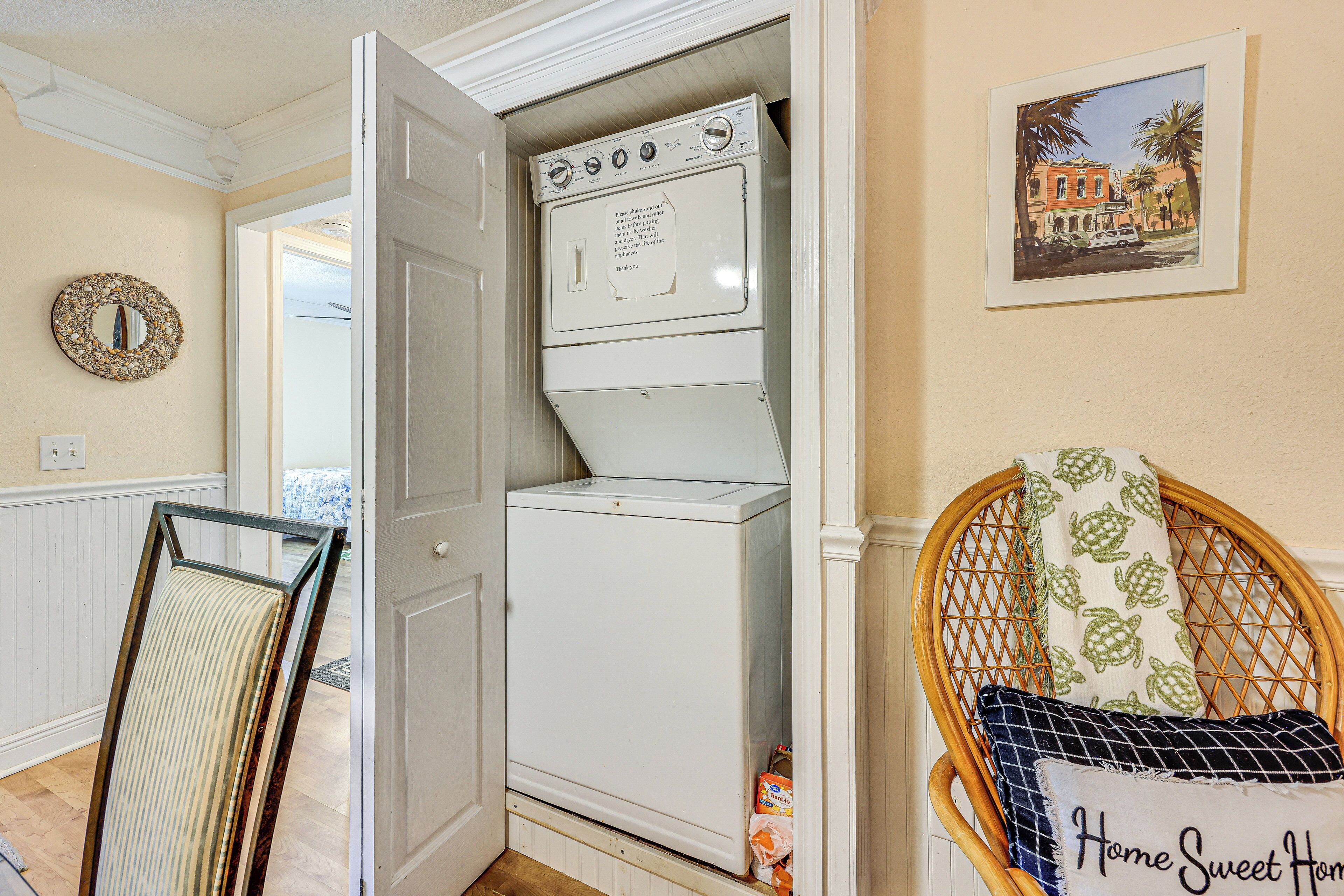 A laundry room with a front-loading washer and dryer, a wicker chair with a cushion, and a framed picture on the wall.