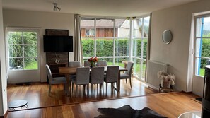 A modern dining area with a wooden table and chairs, a flat-screen TV, and large windows offering a view of a garden and a building.