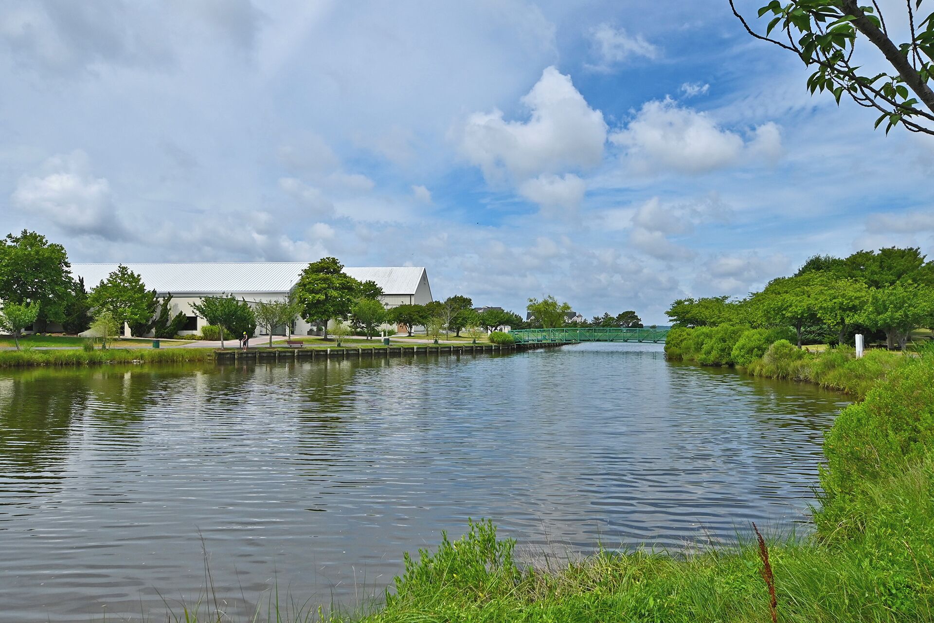 A calm river with a bridge, surrounded by greenery and a building with a white roof.