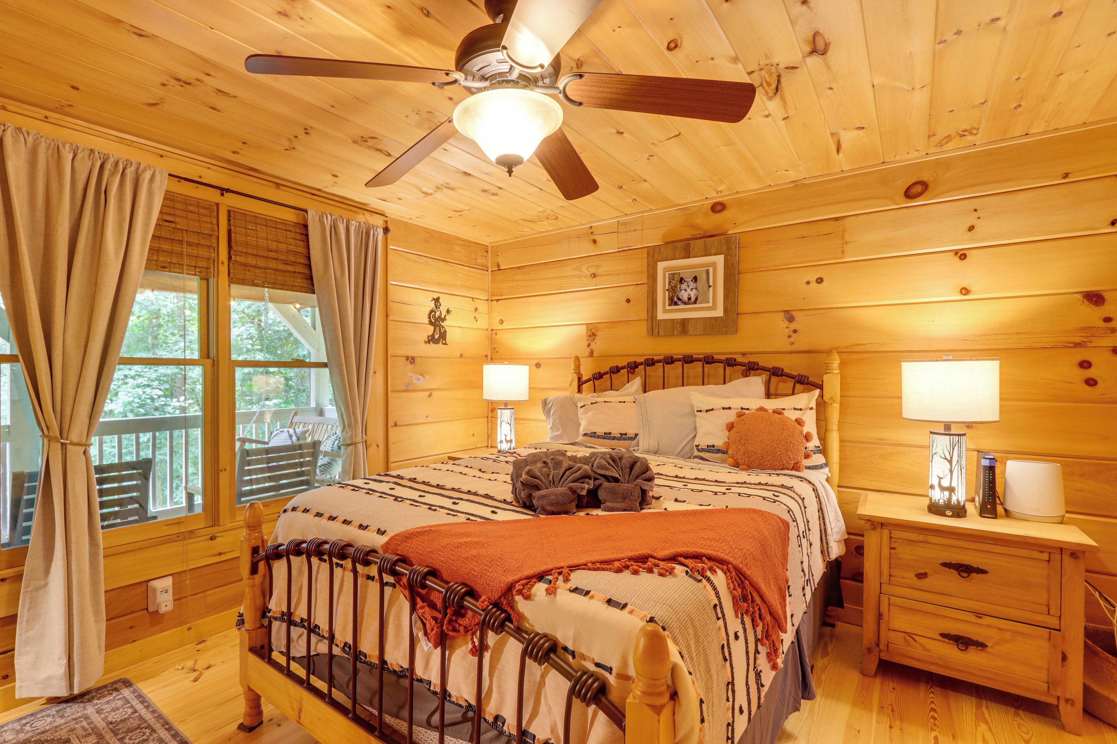 A wooden cabin bedroom with a bed, nightstands, a ceiling fan, and a window with curtains.