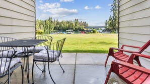 A patio with a table and chairs, a view of a grassy area, and a building in the background. - Waterfront Condo 5-Minute Walk to DT Bigfork (Bigfork)