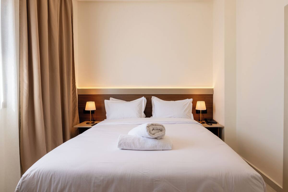 A hotel room with a neatly made bed, two bedside lamps, a wooden headboard, and a curtain on the left side.