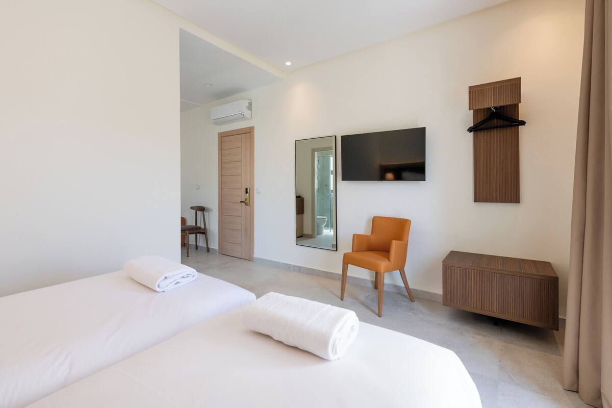 A modern hotel room with two beds, a chair, a mirror, a TV, and a coat rack.