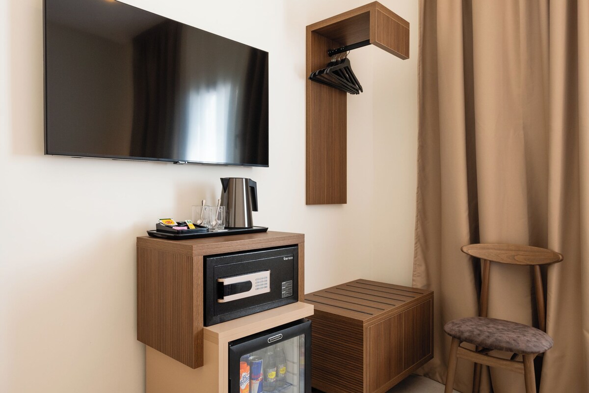 A modern hotel room with a flat-screen TV mounted on the wall, a minibar with a coffee machine, a mini-fridge with beverages, a trash bin, a wooden bench, and a small table with a chair.