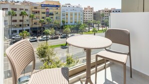 Un balcon avec deux chaises et une table, donnant sur une rue bordée d’immeubles et de palmiers.
