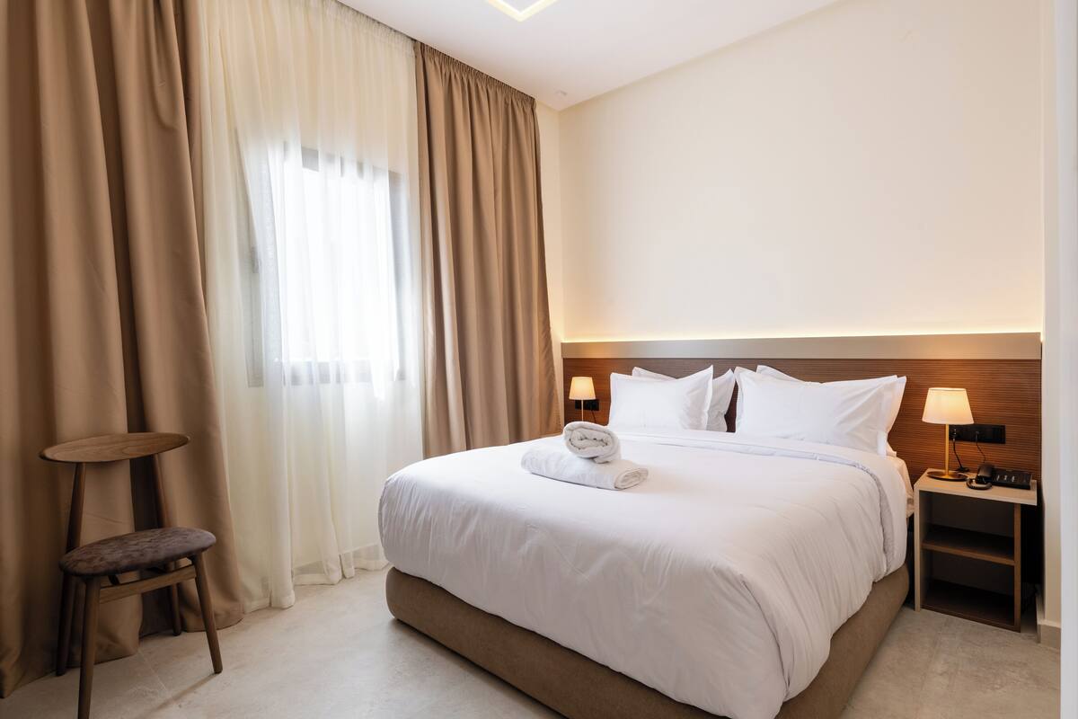 A hotel room with a large bed, a bedside table with a lamp, a chair, and a window with curtains.