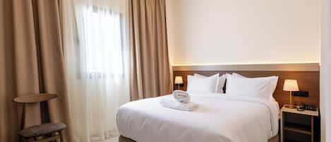 Una habitación de hotel con una cama grande, una mesita de noche con lámpara, una silla y una ventana con cortinas.