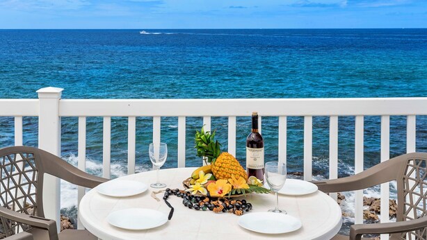 A table set with plates, a bottle of wine, and a fruit arrangement on a balcony overlooking the ocean. - Hale Lani:Direct Oceanfront Massive Pool, Gated, Remodeled Beauty (KAILUA KONA)