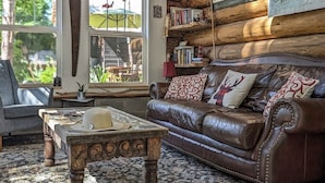 A cozy log cabin interior with a leather sofa, a rustic coffee table, and a bookshelf filled with books and decorative items.