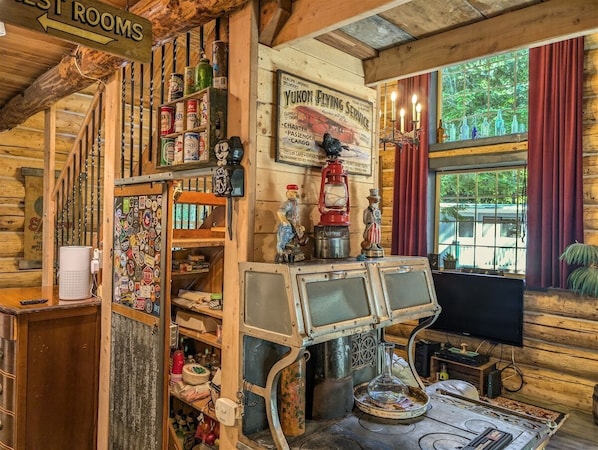 Interior rústico de una cabaña con paredes de madera, un letrero de Yukon Flying Service, una radio antigua y un amplio ventanal con cortinas.