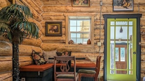 A log cabin interior with a wooden bench, a table with chairs, a green door, and a window with a view.