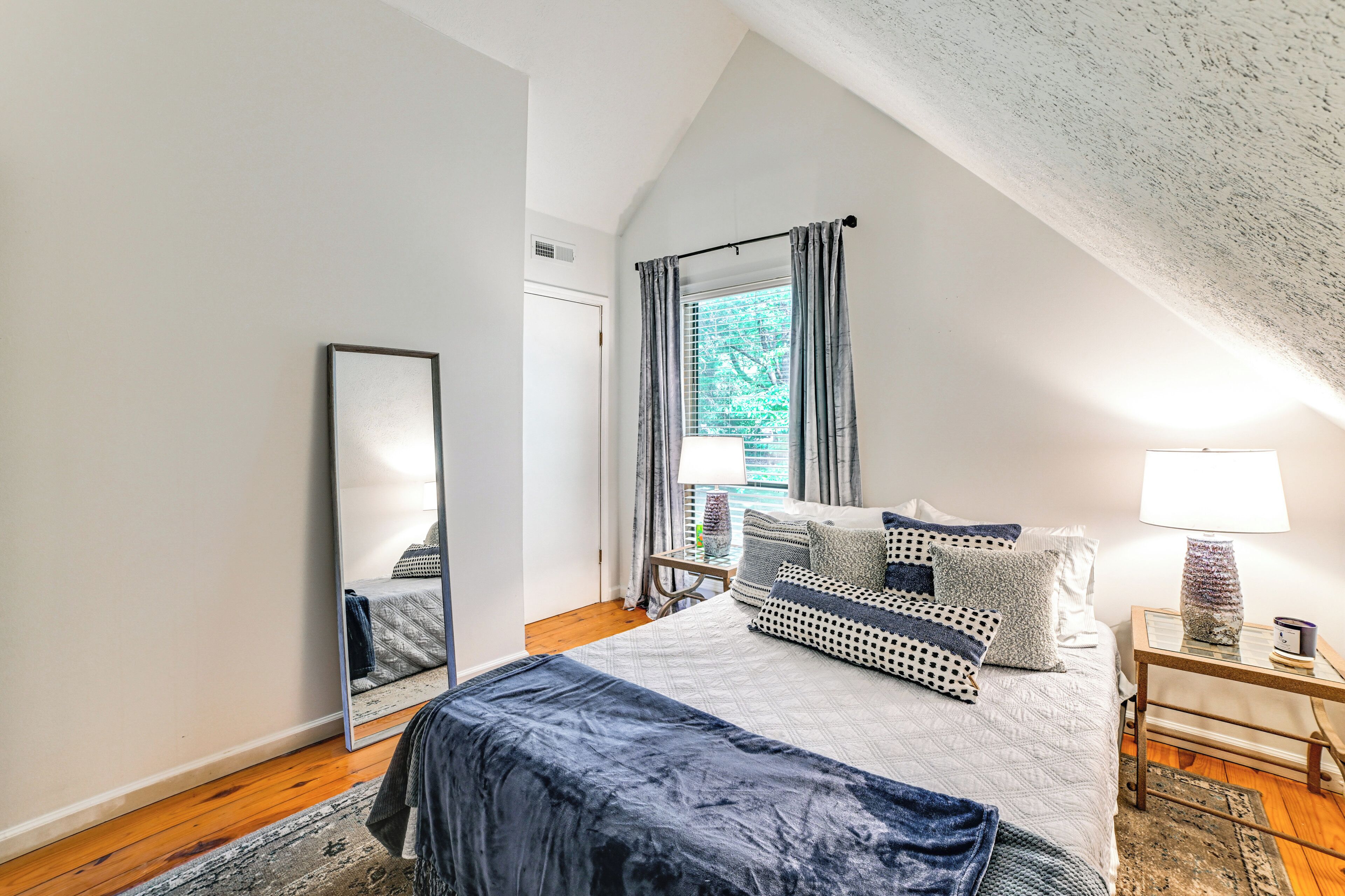 Quarto com uma cama grande, espelho, janela com cortinas, mesa de cabeceira com abajur e um tapete.