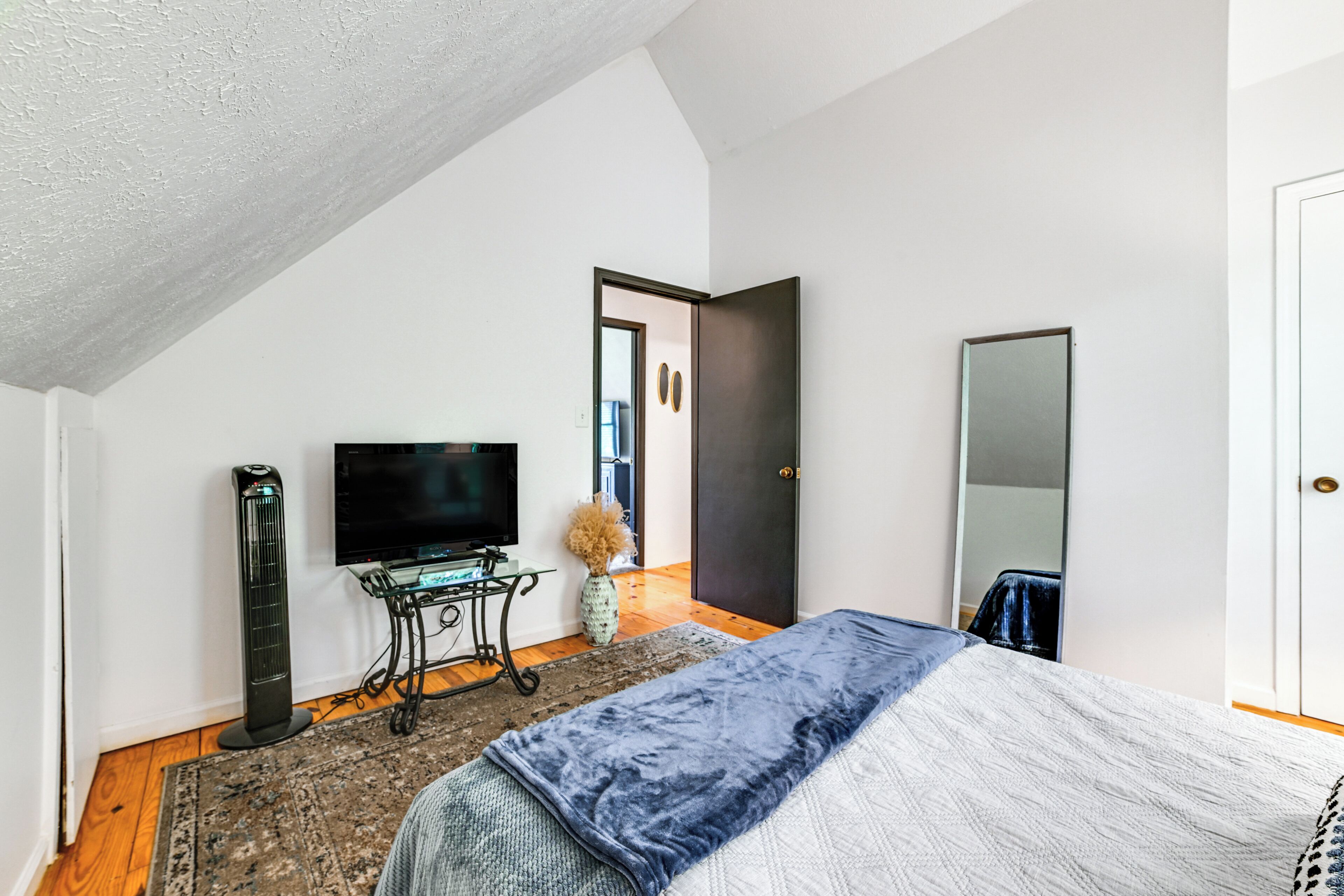 A bedroom with a bed, a TV, a standing fan, and a door leading to another room.