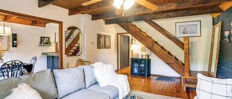Amplia sala de estar con un sofá gris, una mesa de centro y un ventilador de techo. Una escalera con vigas de madera conduce a otra habitación.