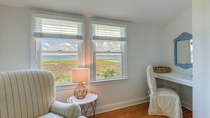 A room with a striped sofa, a small table with a lamp, a chair, and a view of the beach through two windows.