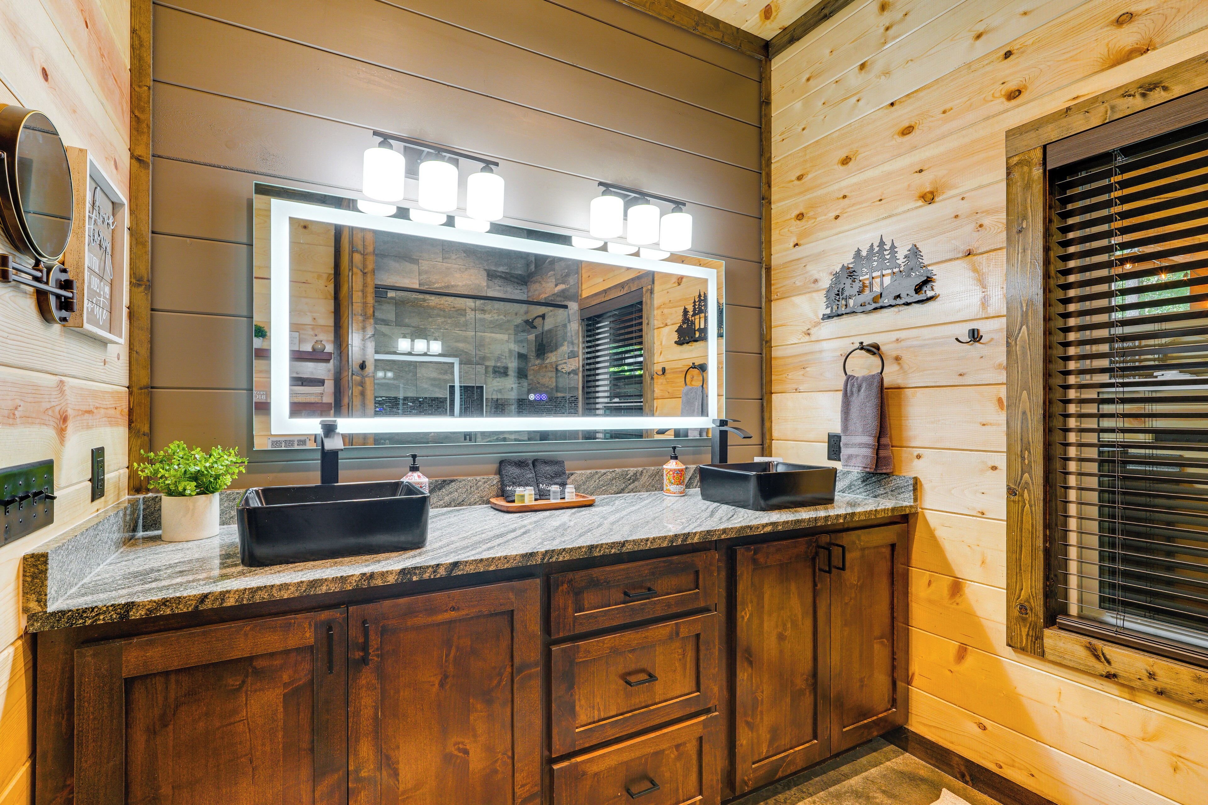 Un bagno in stile baita con elementi in legno, un piano di lavoro in granito, uno specchio grande con illuminazione, un lavabo e mobili impiallacciati.