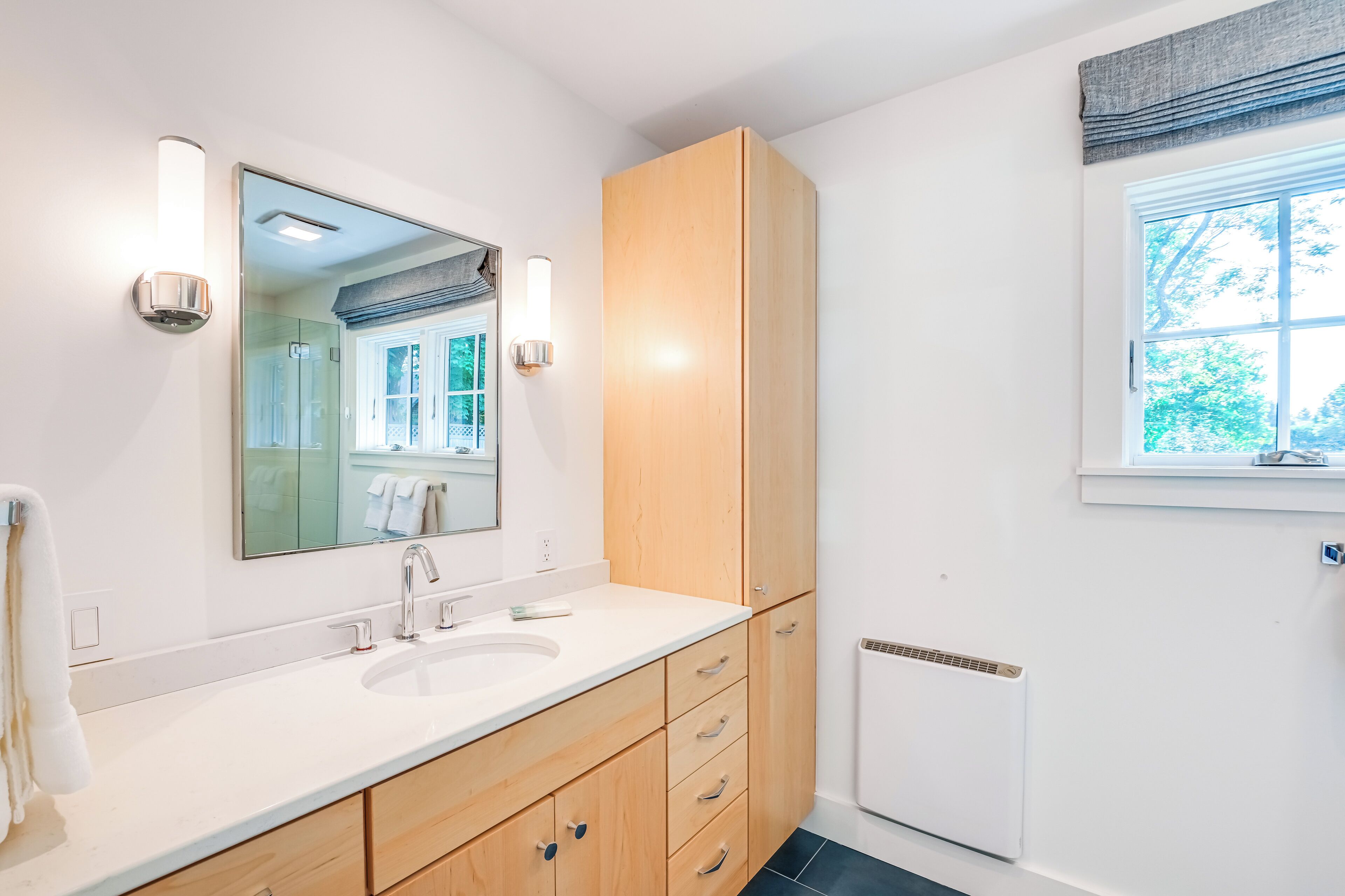 Un baño moderno con un lavamanos blanco, armarios de madera, un espejo grande y un ventanal con vistas a la vegetación.