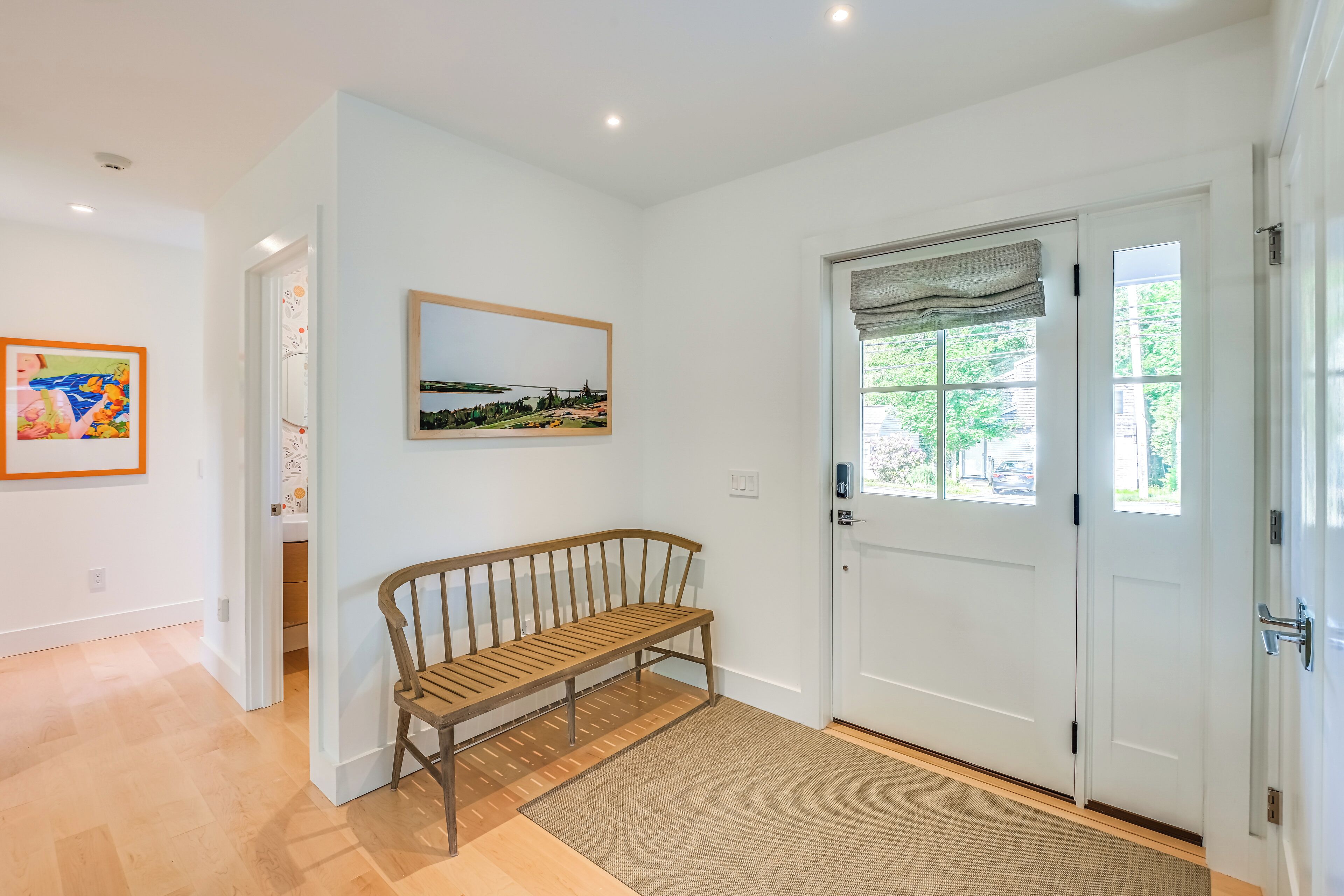 A clean, modern hallway with a wooden bench, a white door with a window, and artwork on the walls.