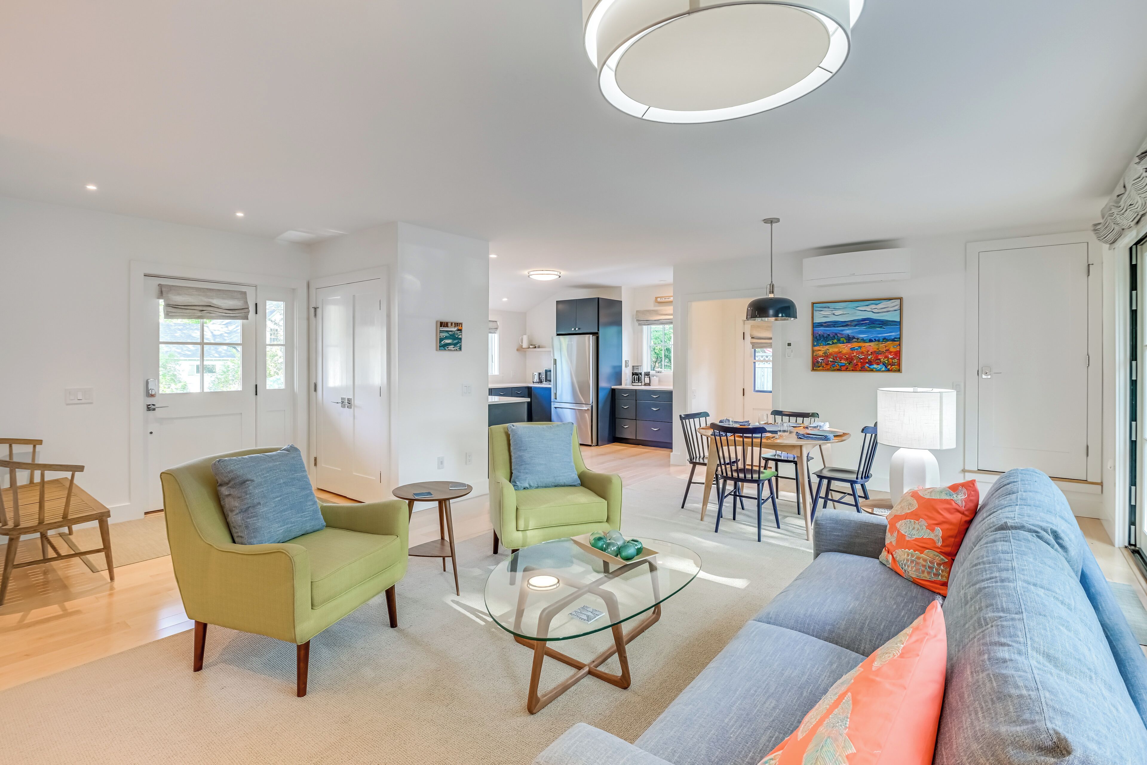 A modern living room with a glass coffee table, two green armchairs, a grey sofa, and a dining area with a table and chairs.