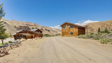 A rustic wooden cabin with a dirt road leading to it, surrounded by mountains and sparse vegetation.