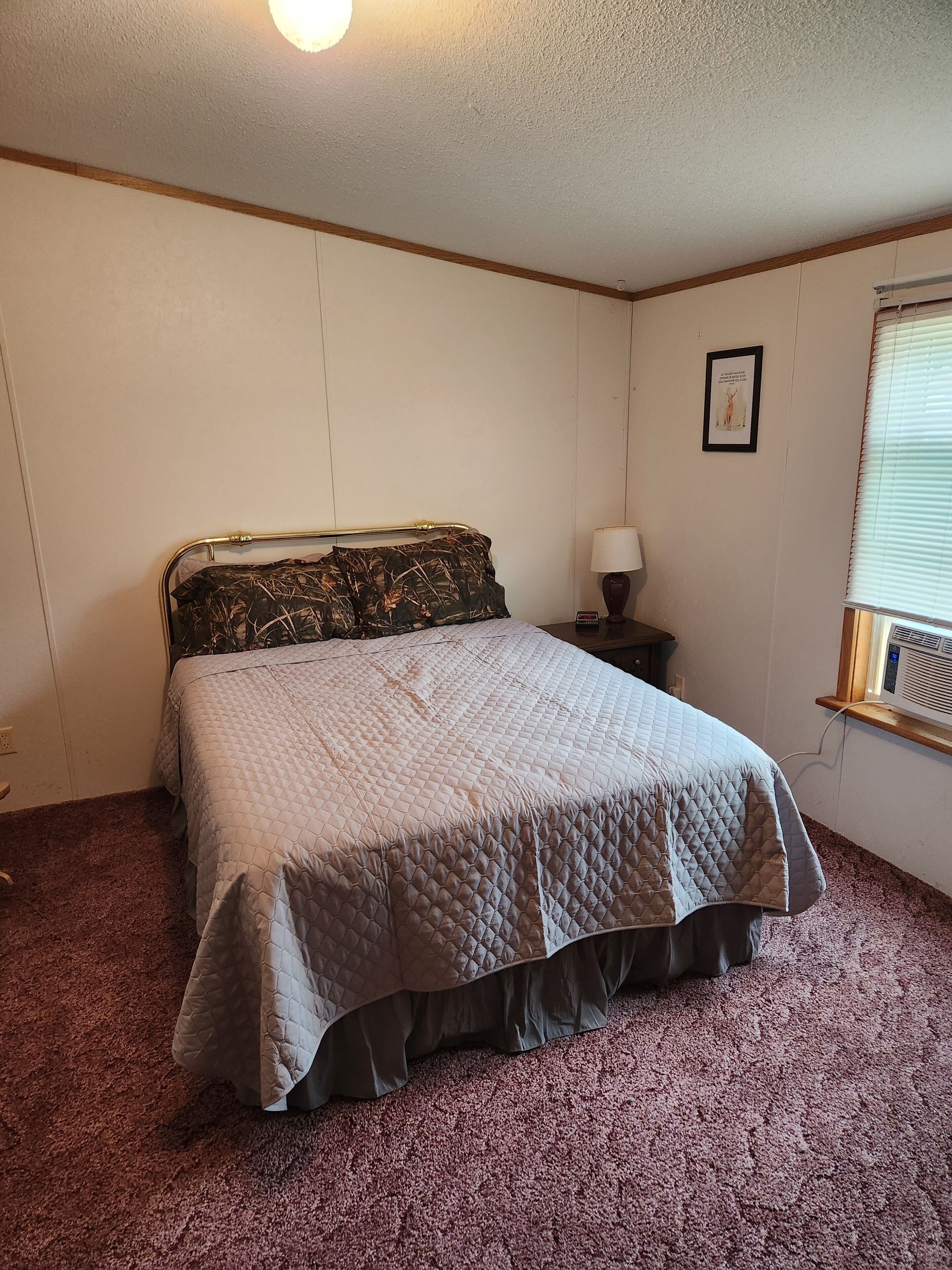 Une chambre à coucher avec un lit, une table de chevet, une lampe, un tableau au mur et une fenêtre avec des stores.