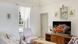 Uma sala de estar com um sofá branco, uma poltrona de couro, uma mesa de centro de madeira e uma televisão sobre um suporte de madeira.
