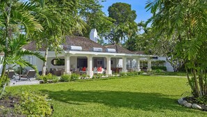 Uma casa de dois andares com varanda coberta, cercada por um gramado bem cuidado e plantas tropicais.