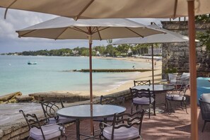 An outdoor seating area with round tables and chairs, overlooking a beach and the sea.