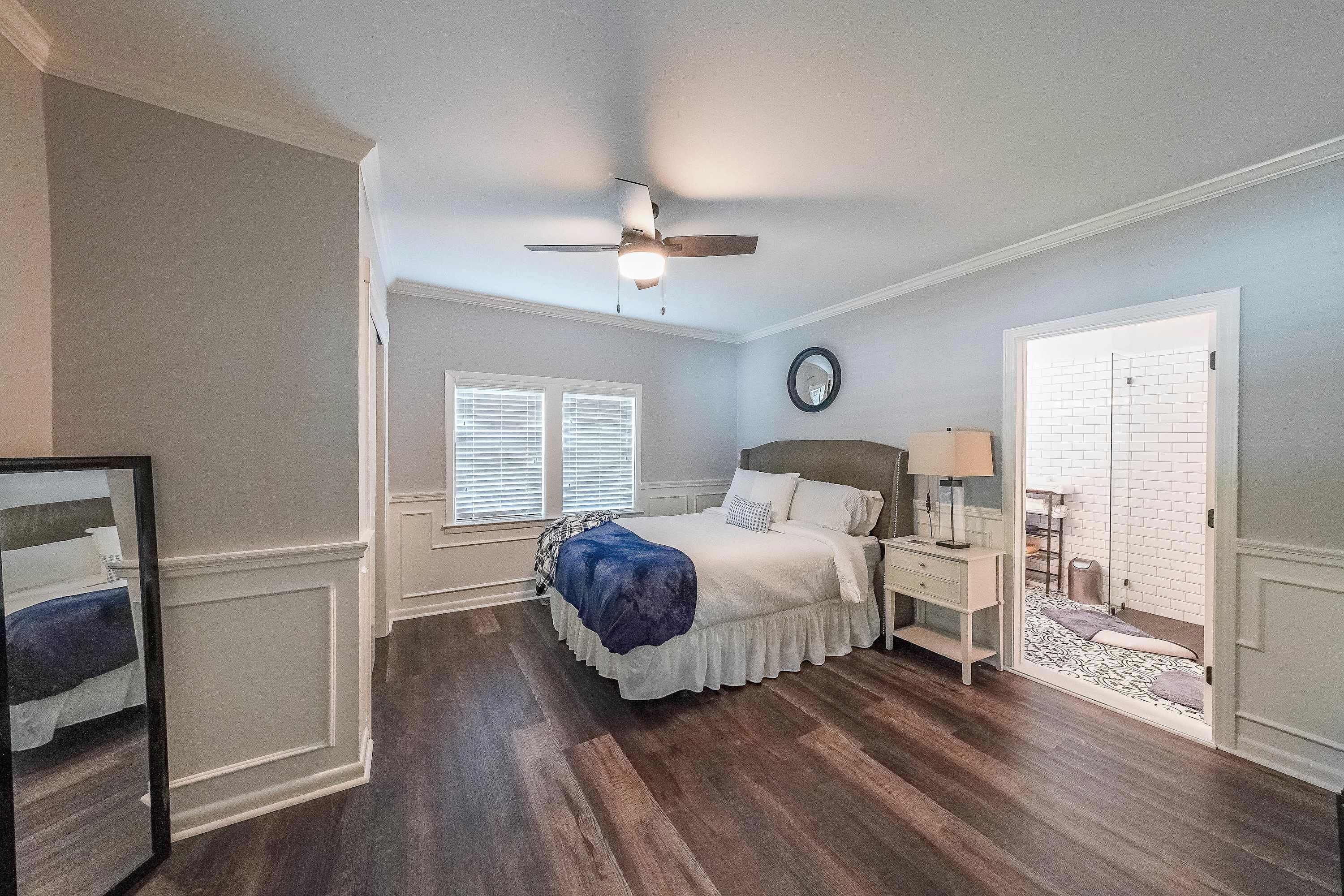 A bedroom with a bed, nightstands, a ceiling fan, a window with blinds, and a door leading to another room.
