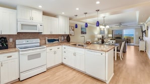 A modern kitchen with white cabinets, a built-in microwave, a stove, and a dishwasher. There is a dining area with a table and chairs, and a view of the ocean through large windows. - 4200 Gulf Drive Unit 108 2 Bedroom Home by RedAwning (Holmes Beach)