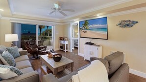 A spacious living room with a large TV, comfortable seating, a ceiling fan, and a view of the beach through the balcony. - 4200 Gulf Drive Unit 108 2 Bedroom Home by RedAwning (Holmes Beach)