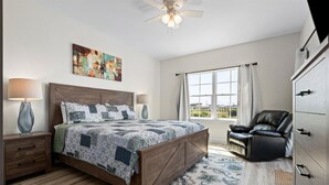 A bedroom with a wooden bed, two bedside lamps, a ceiling fan, a window with curtains, a leather chair, and a dresser. - Sea View Cottage - 110 Ponce De Leon 3 Bedroom Home by RedAwning (Dauphin Island)