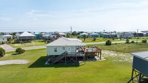 A house with a porch and a small deck, surrounded by a grassy yard and other houses in the background. - Sea View Cottage - 110 Ponce De Leon 3 Bedroom Home by RedAwning (Dauphin Island)
