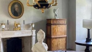 Una habitación con una mesa redonda de madera, una escultura de busto blanco, una chimenea con repisa y una araña.