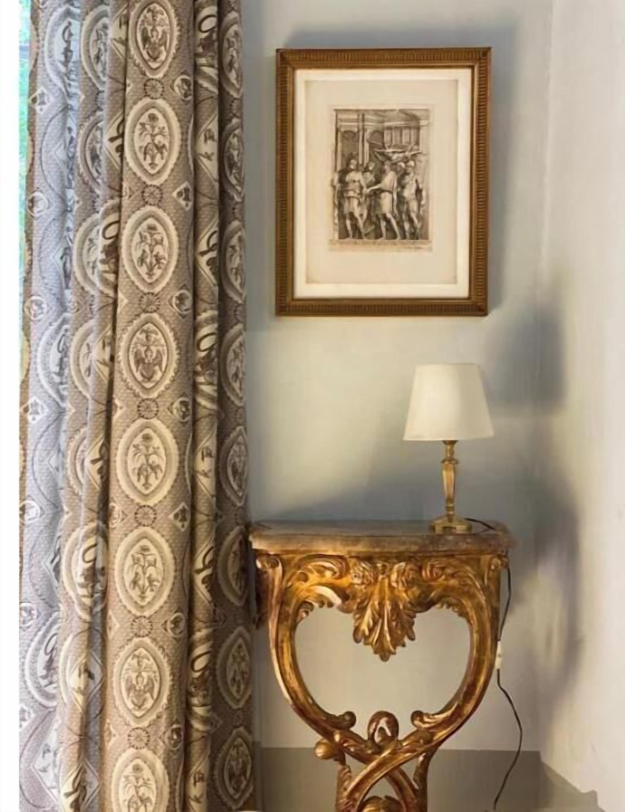 Ein Zimmer mit gemusterten Vorhängen, einem gerahmten Bild an der Wand und einem Tisch mit einer Lampe.