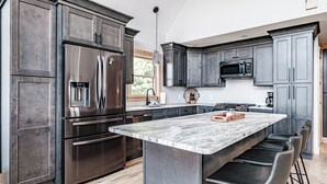 A modern kitchen with dark cabinetry, stainless steel appliances, and a marble island with bar stools. - The Best Mountain Views! You can't stop looking at them! (Hanover)